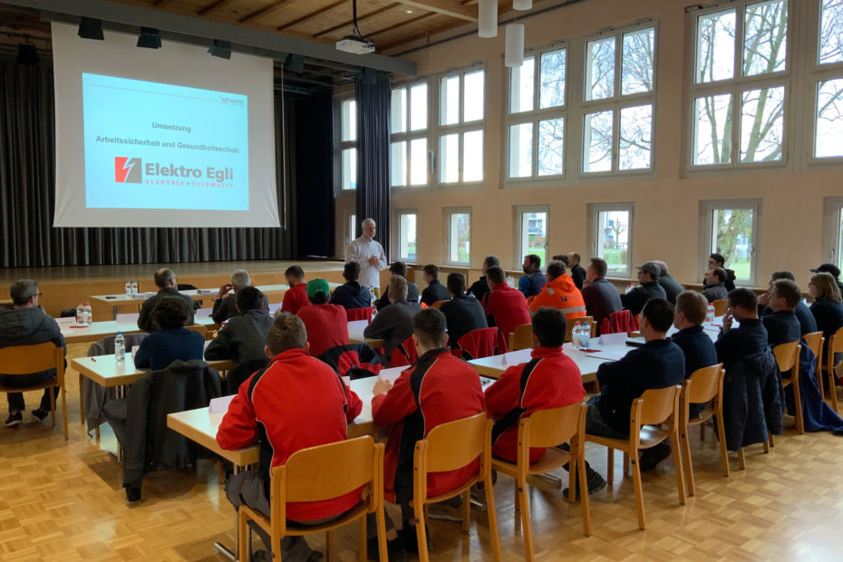 Alle Mitarbeiter sitzen zusammen an Zweiertischen in 3-4 Reihen in einer Aula und schauen einem Referenzen zu